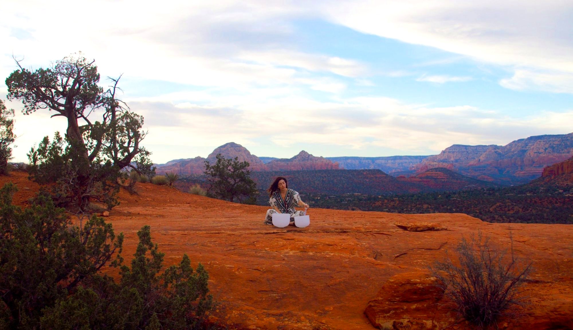 Jennifer Spencer sitting with two singing bowls on the red rock, the Sedona valley beyond